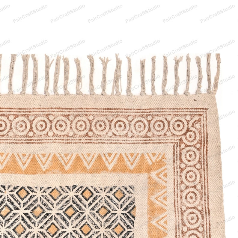 May include: A beige cotton rug with a geometric pattern in black, brown, and yellow. The rug has a fringe border.