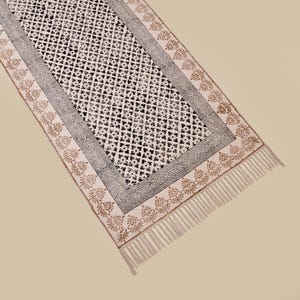 Puede incluir: Una alfombra de pasillo rectangular de color blanquecino con un patr&oacute;n geom&eacute;trico negro repetido en el centro. El borde presenta un dise&ntilde;o negro y gris, con un patr&oacute;n floral marr&oacute;n y flecos en los extremos. La alfombra es de material suave.
