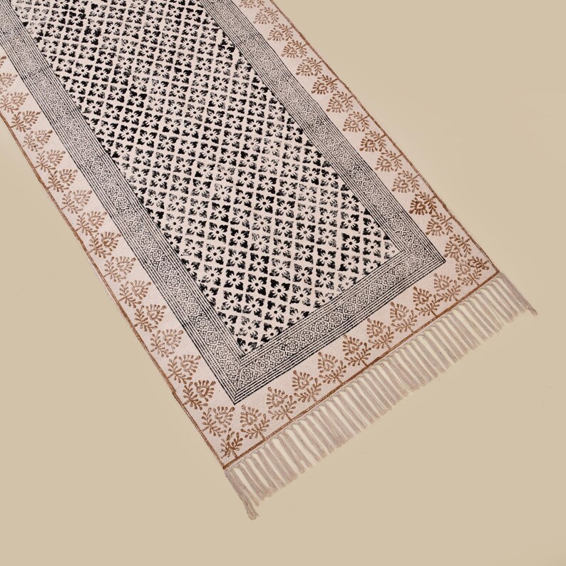 Puede incluir: Una alfombra de pasillo rectangular de color blanquecino con un patr&oacute;n geom&eacute;trico negro repetido en el centro. El borde presenta un dise&ntilde;o negro y gris, con un patr&oacute;n floral marr&oacute;n y flecos en los extremos. La alfombra es de material suave.