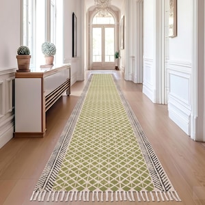 Puede incluir: Una alfombra larga y rectangular con un patrón de diamantes geométricos en tonos verdes y blancos. La alfombra tiene un borde en blanco y negro con flecos en los extremos. La alfombra está colocada en un pasillo con pisos de madera.