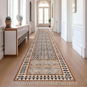 May include: A long, rectangular rug with a geometric pattern in black and cream colors. The rug features a border with a brown trim and fringe at the ends. The rug is placed in a hallway with white walls and wooden flooring.