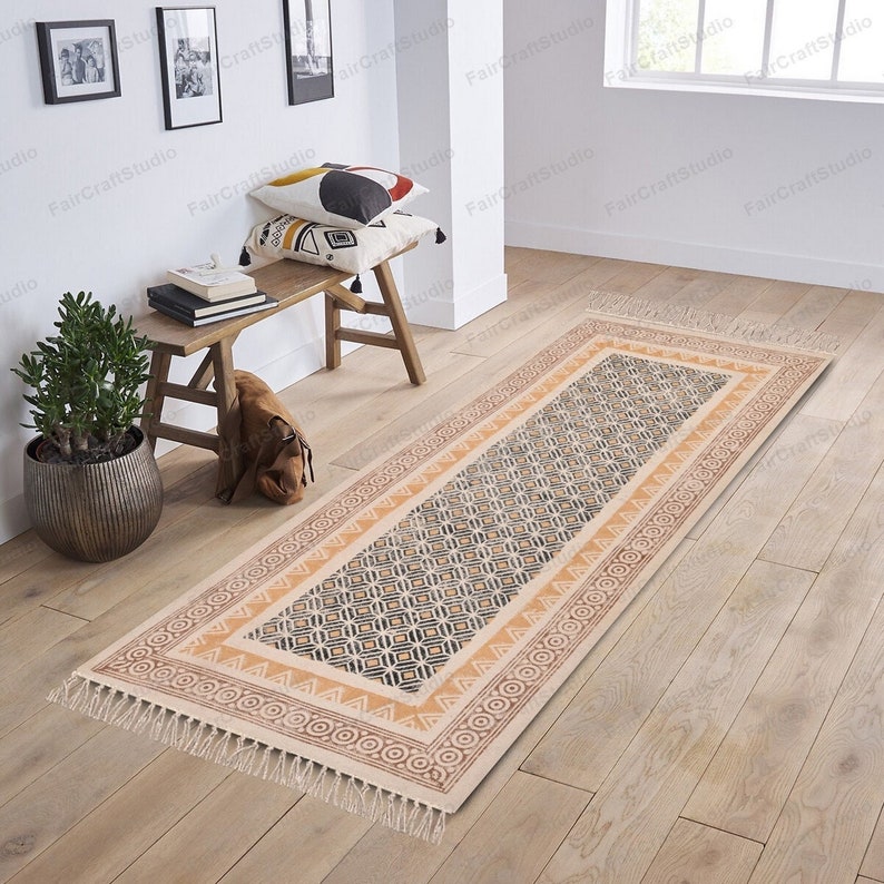 May include: A beige and brown patterned runner rug with a geometric design. The rug has a fringe border and is laid out on a wooden floor.