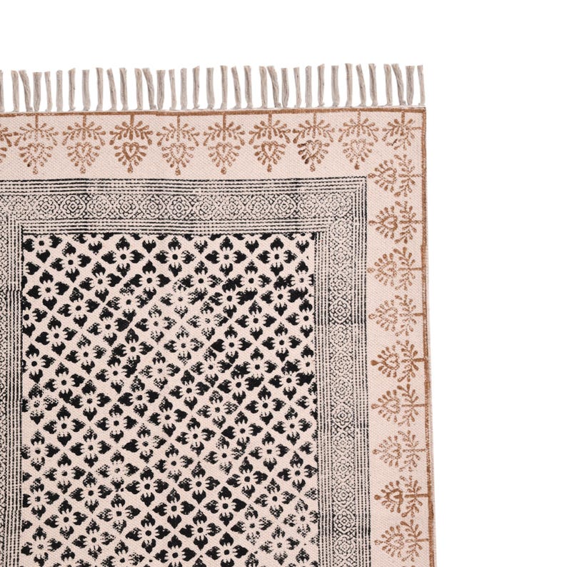 Puede incluir: Alfombra color crema con un estampado floral negro y un borde decorativo. La alfombra tiene flecos en el borde superior y un dise&ntilde;o de hojas repetidas en color marr&oacute;n. Es un art&iacute;culo de decoraci&oacute;n del hogar.