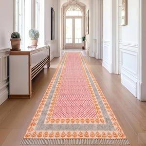 May include: A long, rectangular runner rug with a pink geometric pattern. The rug has a gray border with orange floral designs and fringe at both ends. The rug is placed in a hallway with hardwood floors and white walls.