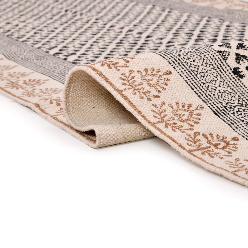 Puede incluir: Una alfombra crema enrollada con un patr&oacute;n geom&eacute;trico. La alfombra presenta dise&ntilde;os en negro y marr&oacute;n, incluyendo un borde con motivos de hojas. La textura de la alfombra es visible y el dise&ntilde;o es sim&eacute;trico.