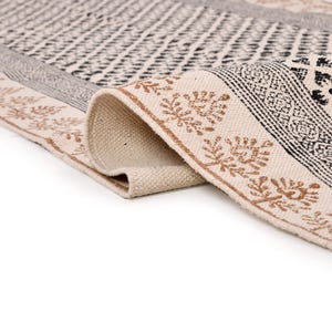 Puede incluir: Una alfombra enrollada de color blanquecino con un patr&oacute;n geom&eacute;trico. La alfombra presenta dise&ntilde;os en negro y marr&oacute;n, incluido un borde con motivos de hojas. La textura de la alfombra es visible y el dise&ntilde;o es sim&eacute;trico.