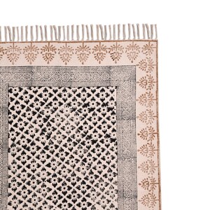 Puede incluir: Alfombra color crema con un estampado floral negro y un borde decorativo. La alfombra tiene flecos en el borde superior y un dise&ntilde;o de hojas repetidas en color marr&oacute;n. Es un art&iacute;culo de decoraci&oacute;n del hogar.