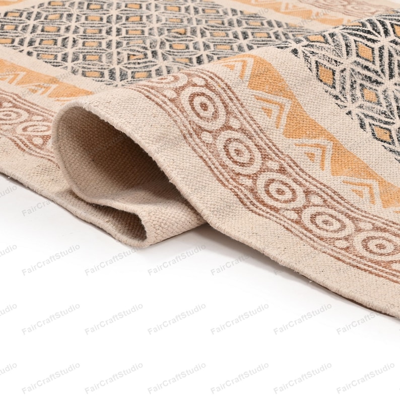 May include: Close-up of a beige cotton rug with a brown and yellow geometric pattern. The rug has a fringed edge and is folded in half.