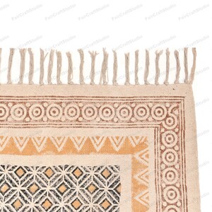 May include: A beige cotton rug with a geometric pattern in black, brown, and yellow. The rug has a fringe border.
