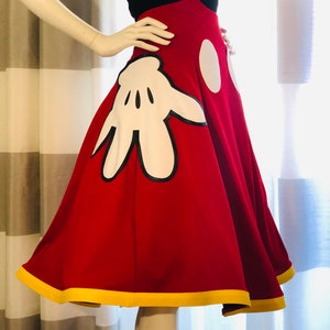 Red high-waisted A-line skirt with yellow trimmed hem, featuring hidden pocket Mickey glove fabric appliqués. Two white vinyl ovals at the center front represent Mickey Mouse's pants.