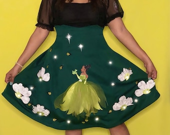Può includere: Un vestito verde con corpetto nero e maniche trasparenti. La gonna presenta un'illustrazione di principessa, fiori bianchi e dettagli scintillanti. L'abito è al ginocchio e abbinato a ballerine scintillanti.