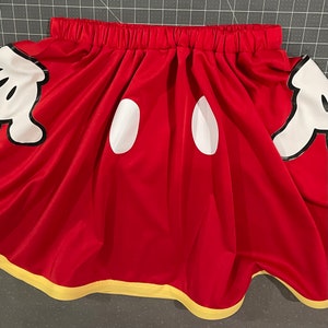 Red elastic waist A-line skirt with yellow trimmed hem, featuring hidden pocket Mickey glove fabric appliqués. Two white vinyl ovals at the center front represent Mickey Mouse's pants.