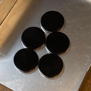 May include: Five black round coasters with a white rim. The coasters are arranged in a pyramid shape on a silver surface.