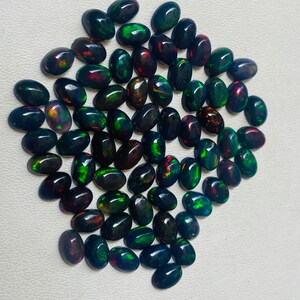 Puede incluir: Una colecci&oacute;n de &oacute;palos negros ovalados con destellos de colores vibrantes, incluyendo verde, rojo y dorado. Las piedras preciosas est&aacute;n dispuestas sobre una superficie blanca, mostrando sus superficies pulidas y reflectantes. Ideales para joyer&iacute;a.