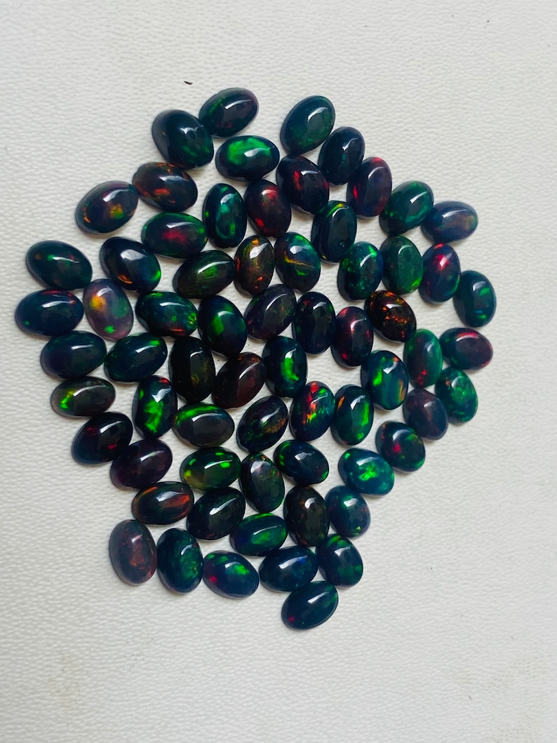 Puede incluir: Una colecci&oacute;n de &oacute;palos negros ovalados con destellos de colores vibrantes, incluyendo verde, rojo y dorado. Las piedras preciosas est&aacute;n dispuestas sobre una superficie blanca, mostrando sus superficies pulidas y reflectantes. Ideales para joyer&iacute;a.
