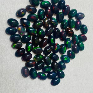 Puede incluir: Una colecci&oacute;n de &oacute;palos negros ovalados con destellos vibrantes de color, incluyendo verde, rojo y azul. Los &oacute;palos est&aacute;n dispuestos muy juntos, mostrando sus superficies pulidas y patrones &uacute;nicos. Las piedras preciosas se exhiben sobre una superficie clara.