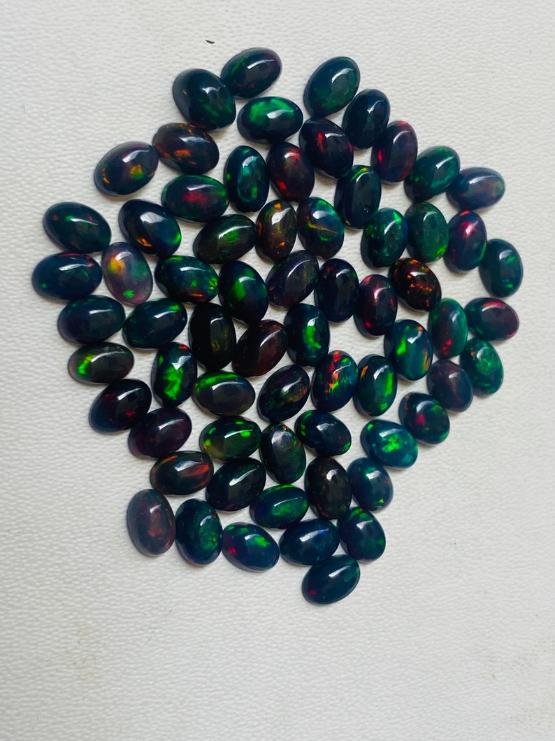 Puede incluir: Una colecci&oacute;n de &oacute;palos negros ovalados con destellos vibrantes de color, incluyendo verde, rojo y azul. Los &oacute;palos est&aacute;n dispuestos muy juntos, mostrando sus superficies pulidas y patrones &uacute;nicos. Las piedras preciosas se exhiben sobre una superficie clara.