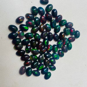 Puede incluir: Una colecci&oacute;n de &oacute;palos negros ovalados con destellos de colores vibrantes, incluyendo verde, rojo y azul. Los &oacute;palos est&aacute;n dispuestos cerca, mostrando sus patrones &uacute;nicos y cualidades iridiscentes. Adecuado para la fabricaci&oacute;n de joyas.