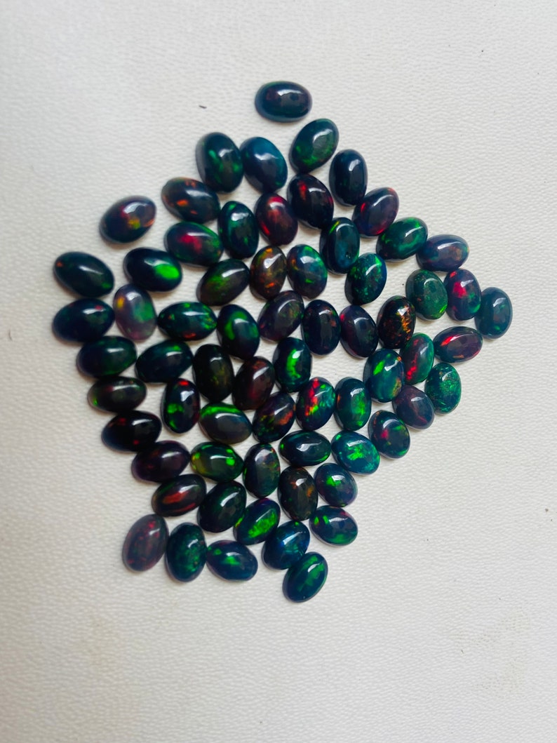 Puede incluir: Una colecci&oacute;n de &oacute;palos negros ovalados con destellos de colores vibrantes, incluyendo verde, rojo y azul. Los &oacute;palos est&aacute;n dispuestos cerca, mostrando sus patrones &uacute;nicos y cualidades iridiscentes. Adecuado para la fabricaci&oacute;n de joyas.