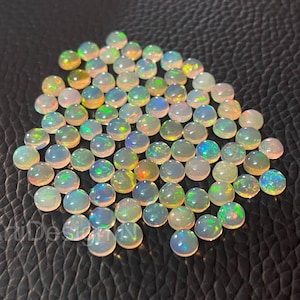 May include: A scattering of small, polished opals, each with a rounded shape. The opals exhibit a play of colour, with flashes of blue, green, and orange within a white or yellow base. The gemstones are arranged on a dark, textured background.