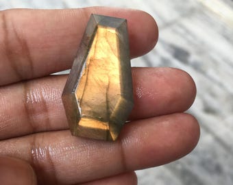 May include: A coffin-shaped labradorite gemstone featuring a faceted surface with a prominent golden-yellow flash. The stone displays a smooth, polished finish with dark grey and brown tones visible along the edges, showcasing natural mineral luster.