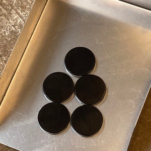 May include: A set of six black round magnetic discs arranged in a pyramid shape on a silver metal surface.
