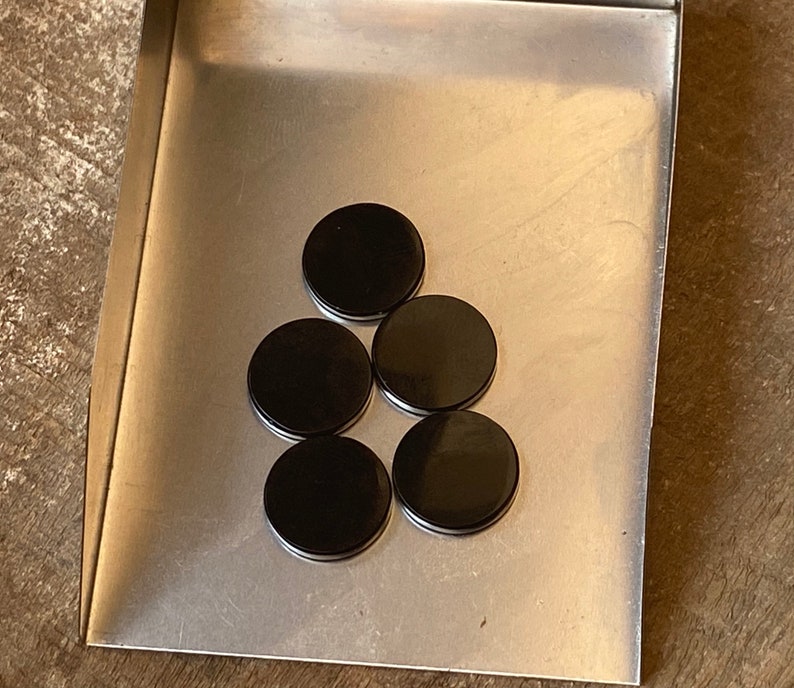 May include: Five black round disc magnets on a silver metal surface.
