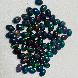 Puede incluir: Una colecci&oacute;n de &oacute;palos negros de forma ovalada con destellos vibrantes de verde, rojo y dorado. Las piedras preciosas est&aacute;n dispuestas sobre una superficie blanca, mostrando sus colores iridiscentes. Ideales para la creaci&oacute;n de joyas.