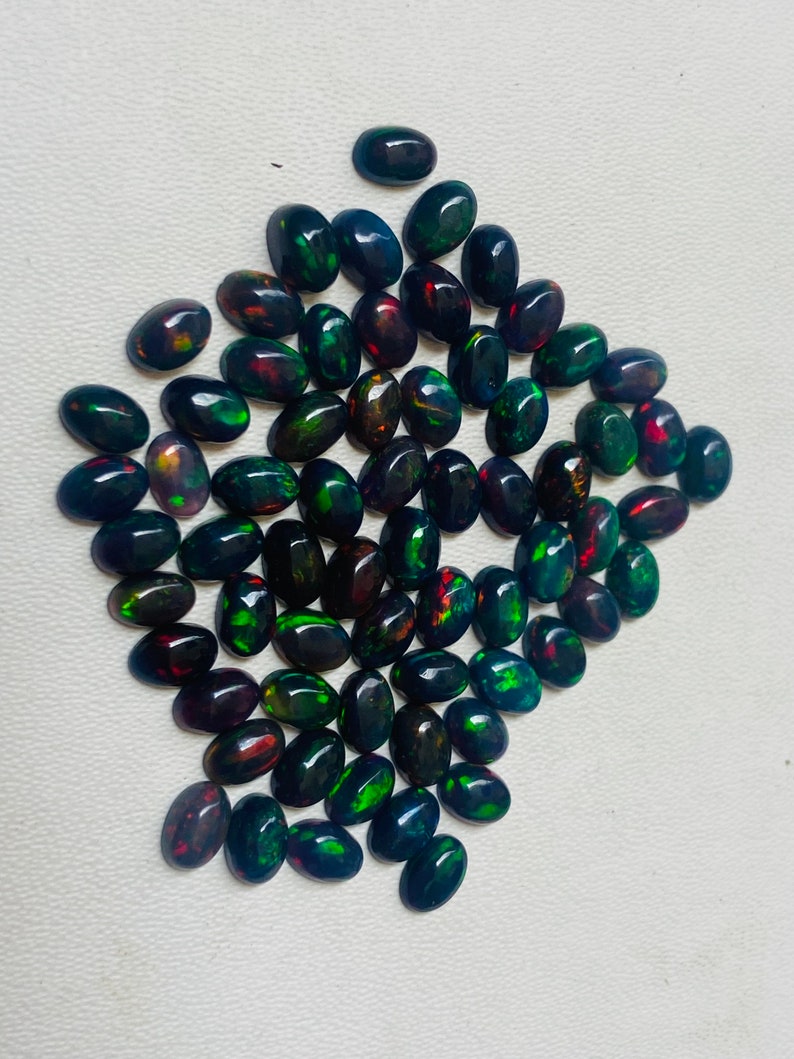 Puede incluir: Una colecci&oacute;n de &oacute;palos negros de forma ovalada con destellos vibrantes de verde, rojo y dorado. Las piedras preciosas est&aacute;n dispuestas sobre una superficie blanca, mostrando sus colores iridiscentes. Ideales para la creaci&oacute;n de joyas.