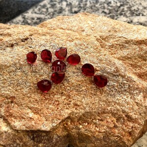 Può includere: Un gruppo di otto pietre preziose rosse a forma di goccia su una superficie rocciosa marrone ruvida.