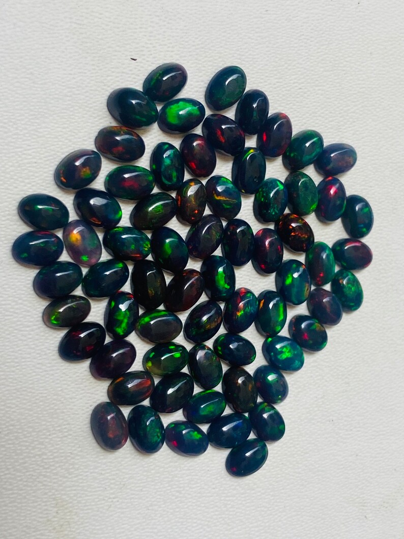 Puede incluir: Un conjunto de &oacute;palos negros de forma ovalada con vibrantes destellos de color, incluyendo verde, rojo y dorado. Las piedras preciosas est&aacute;n dispuestas juntas, mostrando sus superficies pulidas y su juego de colores iridiscente. Ideales para joyer&iacute;a.