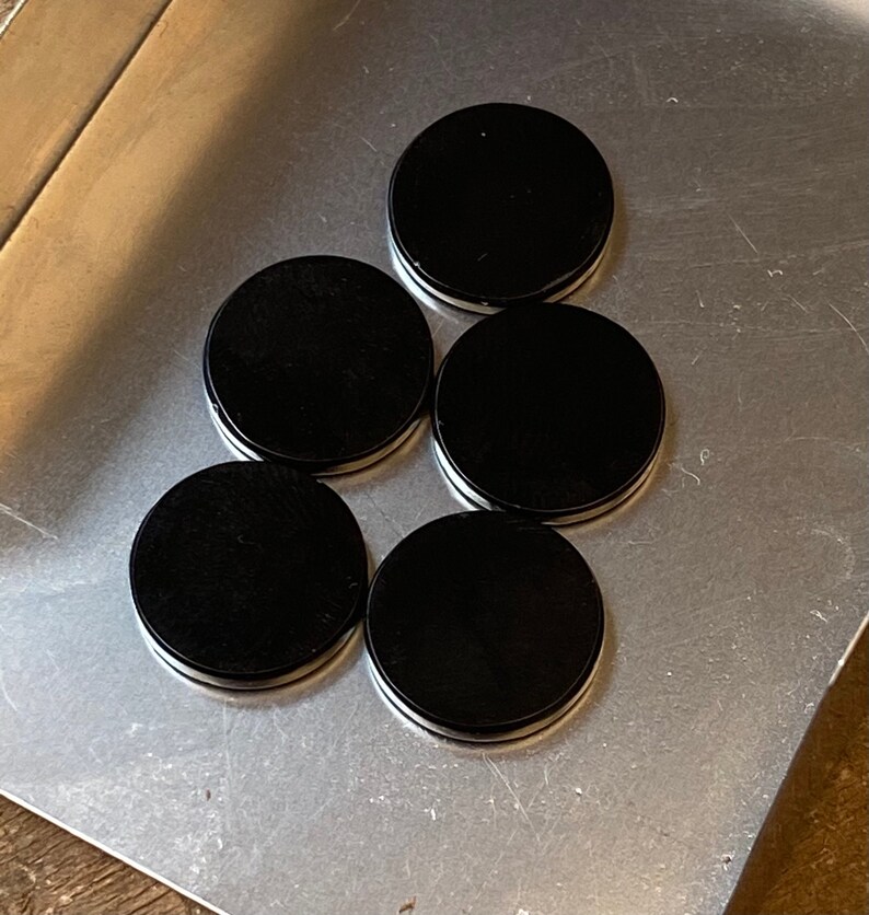May include: Five black round coasters with a white rim. The coasters are arranged in a pyramid shape on a silver surface.