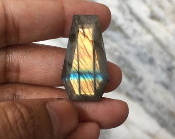May include: A faceted, coffin-shaped labradorite gemstone. The stone displays a play of color with hues of gold, blue, and purple. The gemstone is held in a hand, showcasing its polished surface and unique shape. This gemstone is ideal for jewelry making.