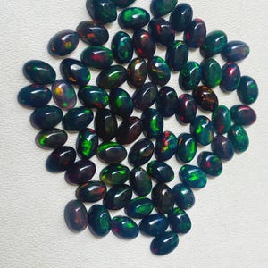 Puede incluir: Una colecci&oacute;n de &oacute;palos negros ovalados con vibrantes destellos verdes, rojos y azules. Las piedras preciosas est&aacute;n dispersas sobre una superficie blanca, mostrando su apariencia pulida y brillante. Estos &oacute;palos son ideales para la fabricaci&oacute;n de joyas.