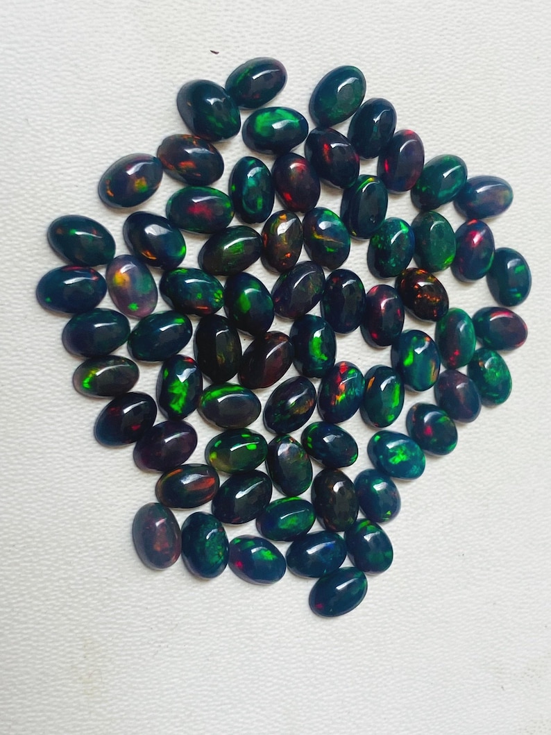 Puede incluir: Una colecci&oacute;n de &oacute;palos negros ovalados con vibrantes destellos verdes, rojos y azules. Las piedras preciosas est&aacute;n dispersas sobre una superficie blanca, mostrando su apariencia pulida y brillante. Estos &oacute;palos son ideales para la fabricaci&oacute;n de joyas.