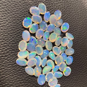 Puede incluir: Una colección de piedras preciosas de ópalo blanco de forma ovalada con colores arcoíris iridiscentes. Los ópalos están dispuestos en un montón suelto sobre una superficie negra texturizada.