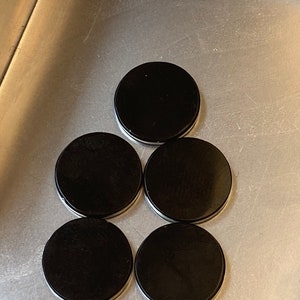 May include: Five black round coasters arranged in a pyramid shape on a silver surface.