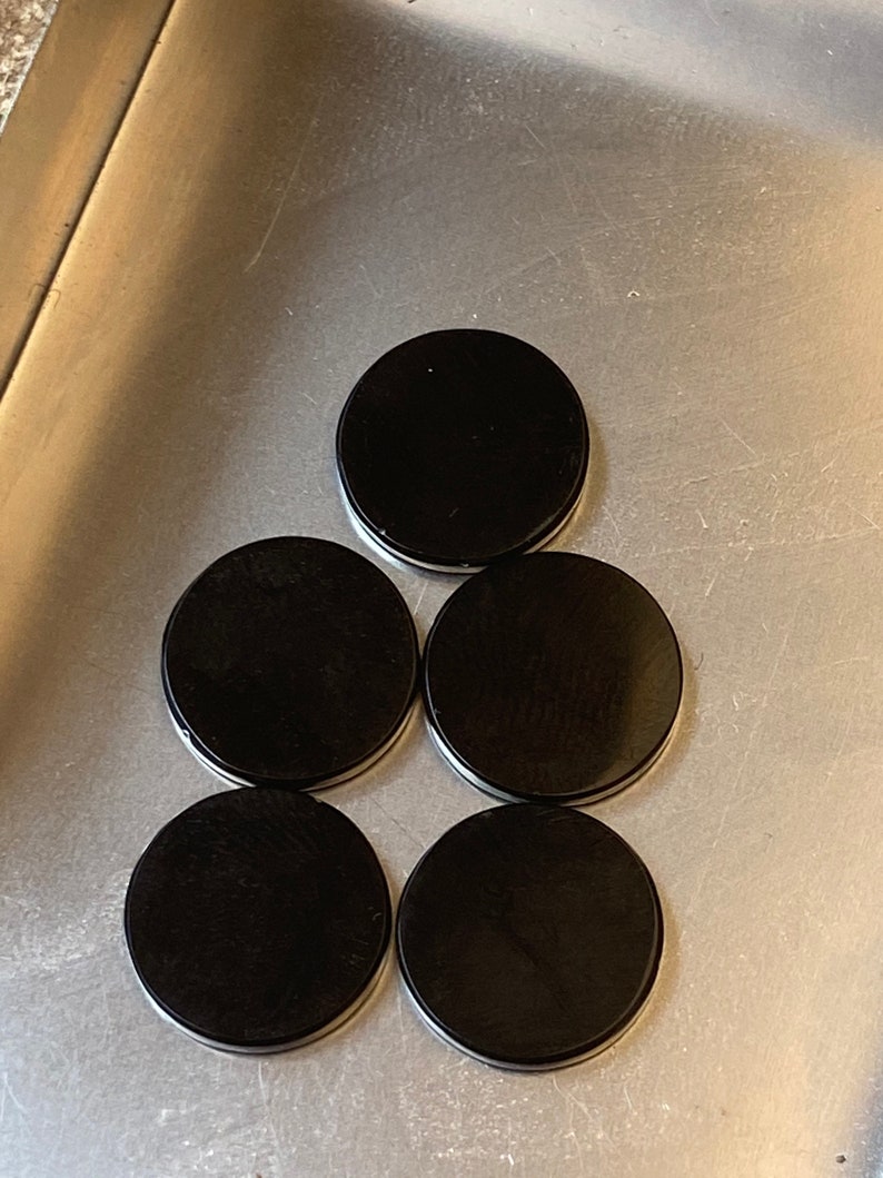 May include: Five black round coasters arranged in a pyramid shape on a silver surface.