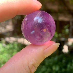May include: A small, polished, spherical amethyst gemstone with a translucent purple hue. The stone features natural internal inclusions and variations in colour intensity throughout its rounded, smooth surface, catching the light.