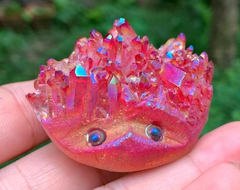 Puede incluir: Un vibrante grupo de cristales rojos con reflejos iridiscentes. El cristal se sostiene en una mano, mostrando su forma única y variaciones de color. El cristal tiene una base redondeada con dos pequeños acentos redondos azul-verdosos.