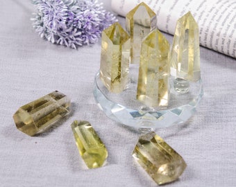 May include: A group of seven clear, yellow citrine crystal points on a clear glass stand. The crystals are various sizes and shapes, with some having a more pointed tip than others.