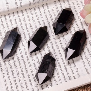 Puede incluir: Seis puntas de cristal de obsidiana negra, también conocidas como cuarzo negro, están dispuestas en una página de un libro. Los cristales son puntiagudos en ambos extremos y tienen una superficie lisa y pulida.
