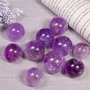 May include: A collection of ten amethyst crystal spheres of varying sizes, ranging in colour from light to dark purple, displayed on a textured, light grey surface.