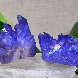 May include: Two clusters of iridescent blue quartz crystals on a white surface. The crystals are arranged in a cluster and have a variety of shapes and sizes.