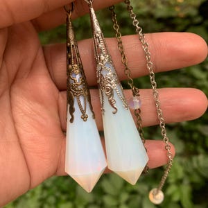 Puede incluir: Dos péndulos de ópalo blanco con engastes ornamentados de plata y bronce que cuelgan de cadenas de plata y bronce.