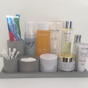 May include: A grey concrete tray with various skincare and beauty products, including a tube of toothpaste, a bottle of hand cream, a bottle of body oil, and a jar of face cream. The products are arranged neatly and tidily on the tray.