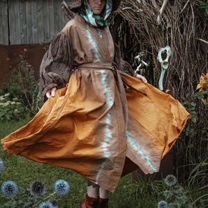 May include: A brown and orange tie-dye robe with a matching hat. The robe has long sleeves and a belt. The person is wearing red boots. The background includes a wooden fence and greenery.