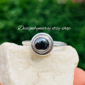 Pode incluir: Um anel de prata com uma pedra de hematita preta engastada em um engaste de bezel. O anel está sobre uma rocha branca.