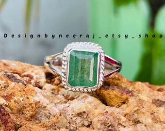 Puede incluir: Un anillo de plata con una piedra preciosa rectangular de color verde esmeralda. La piedra preciosa está engastada en un borde decorativo en forma de cuerda. El anillo se muestra sobre una roca marrón texturizada. El texto "Designbyneeraj_etsy_shop" es visible.