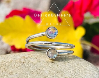 May include: A silver ring with two iridescent moonstone gems. The ring is a double band, with one band wrapping around the other. The ring is on a gray stone background.